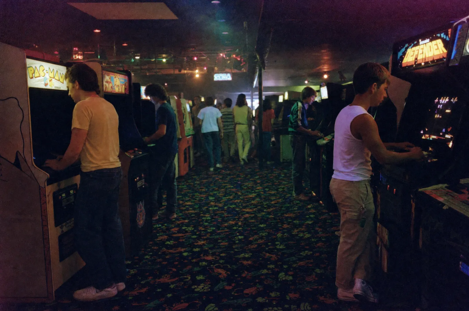 The interior of a 1985 video arcade generated from prompt 48: cabinet screens and marquees lighting players' faces, mixed uncorrected colour temperatures, heavy pushed-film grain and patterned carpet